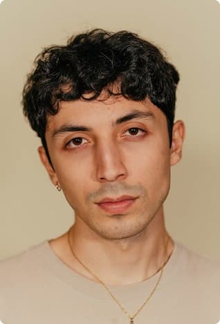 Portrait of a young man with dark curly hair and a thin necklace, warm backdrop.