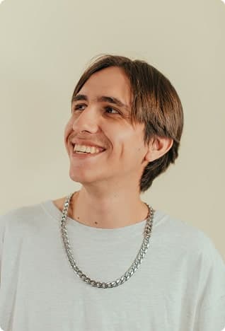 Man with medium-length hair, a chain necklace and light background, slight smile.
